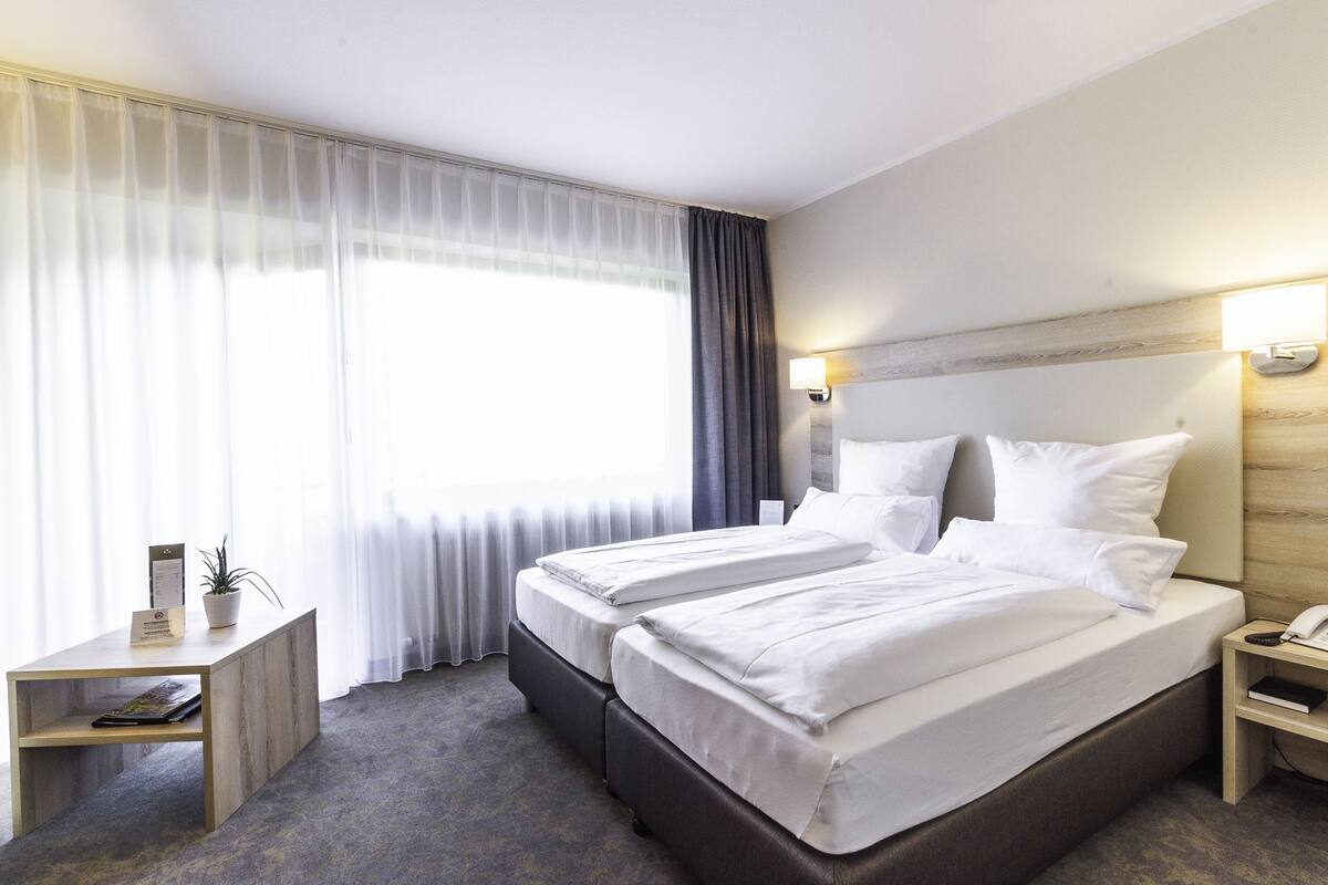 Ein Hotelzimmer mit Doppelbett, Nachttisch, Lampe und Blick ins Freie durch transparente Vorhänge.