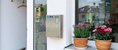 Une sonnette dotée d’un clavier et de différentes options de paiement, une fenêtre donnant sur un immeuble, et deux plantes en pot posées sur le rebord de la fenêtre.