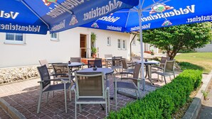 An outdoor patio with tables and chairs, blue umbrellas, and a white building in the background. - Motel Hormersdorf (Schnaittach)