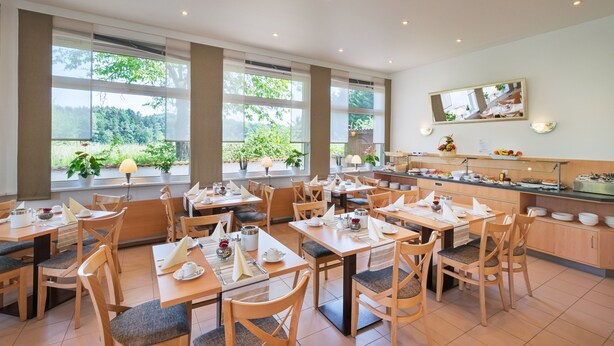 A spacious dining area with multiple tables and chairs, large windows with a view of greenery, and a buffet counter with various food items. - Motel Hormersdorf (Schnaittach)