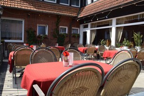 Une terrasse extérieure avec des chaises et des tables en osier, dont certaines sont recouvertes de nappes rouges, et des plantes en pot.