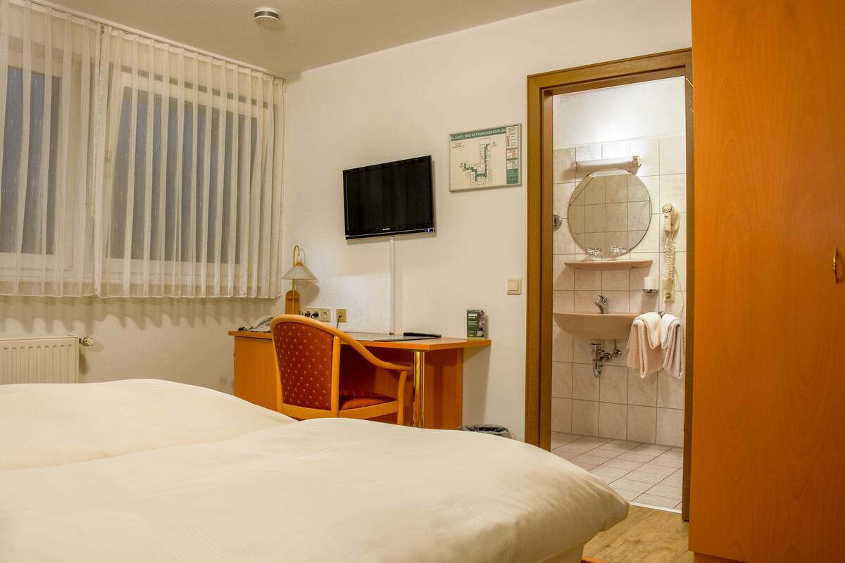 A hotel room with a bed, a desk with a chair, a television, a bathroom with a sink and mirror, and a door leading to the bathroom.