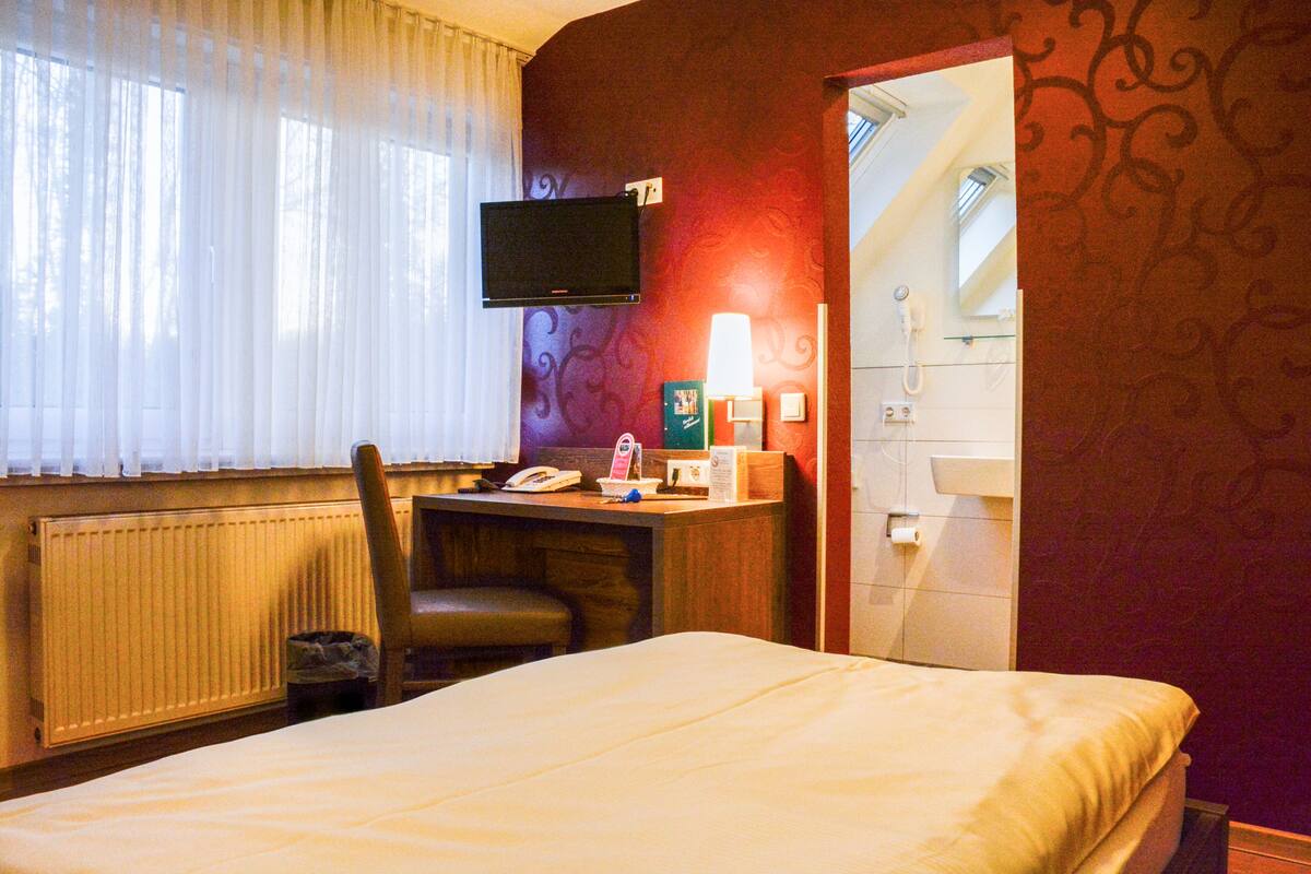 A hotel room with a bed, a desk with a chair, a television mounted on the wall, a lamp, and a bathroom visible through an open door.