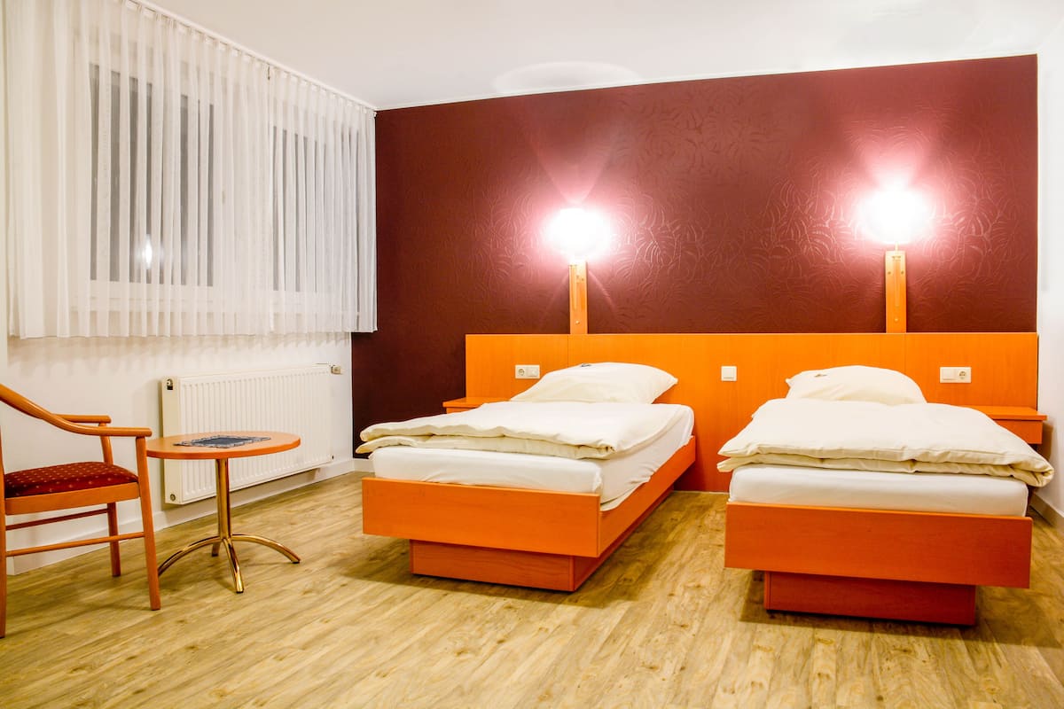 A hotel room with two beds, a chair, a small table, and a radiator.