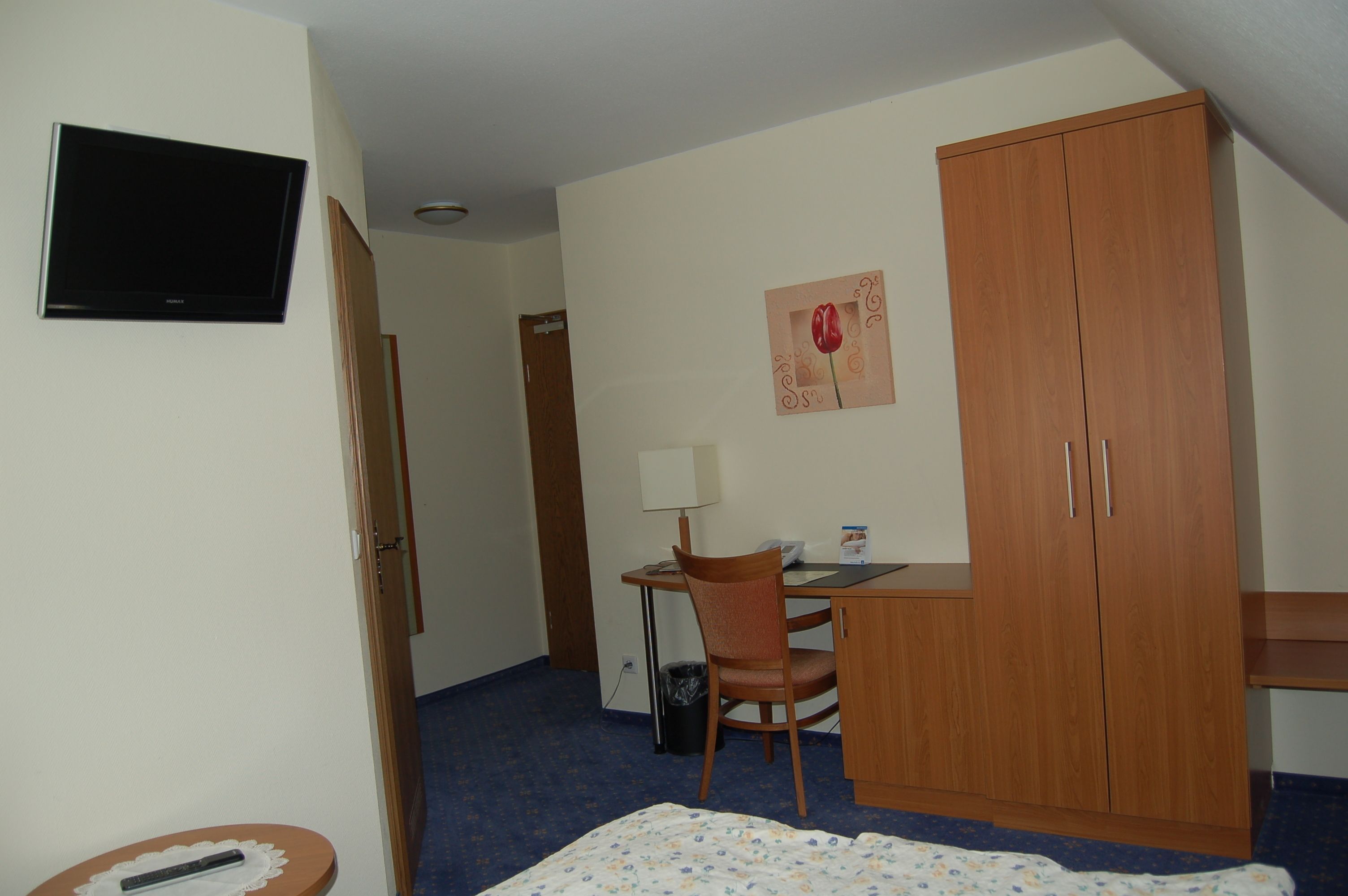 A hotel room with a bed, a desk with a chair, a television, a wardrobe, and a painting on the wall.
