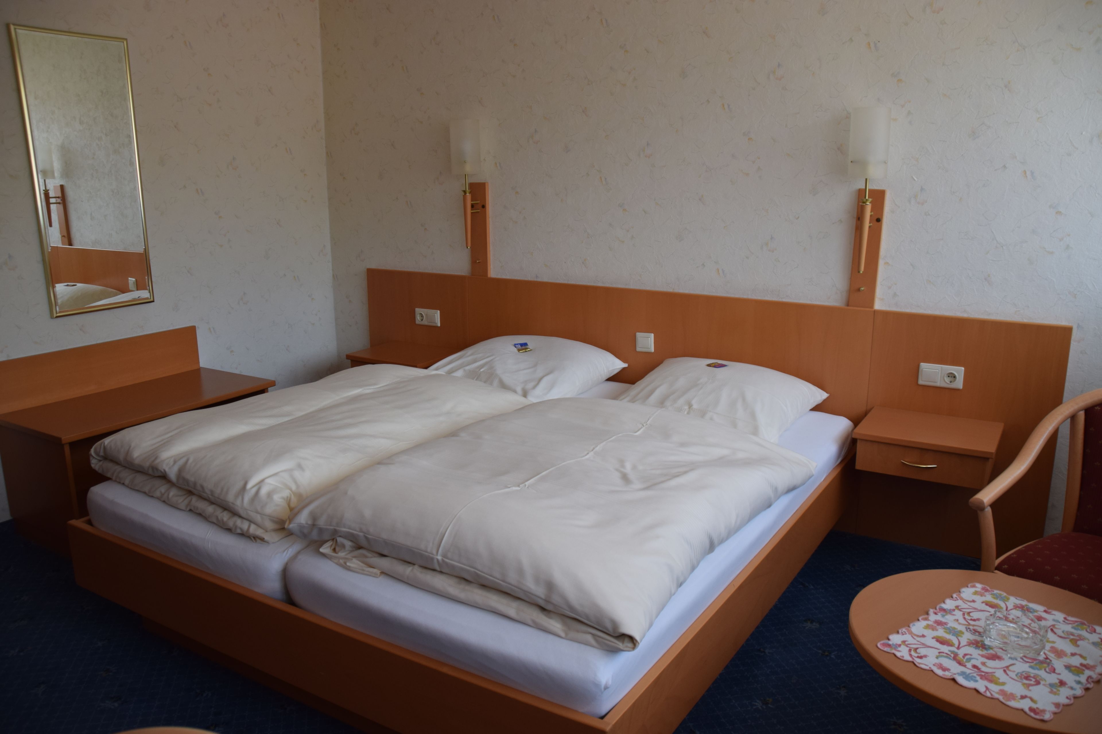 Habitación de hotel con cama de madera, dos mesitas de noche, una silla y una pequeña mesa redonda con mantel floreado.