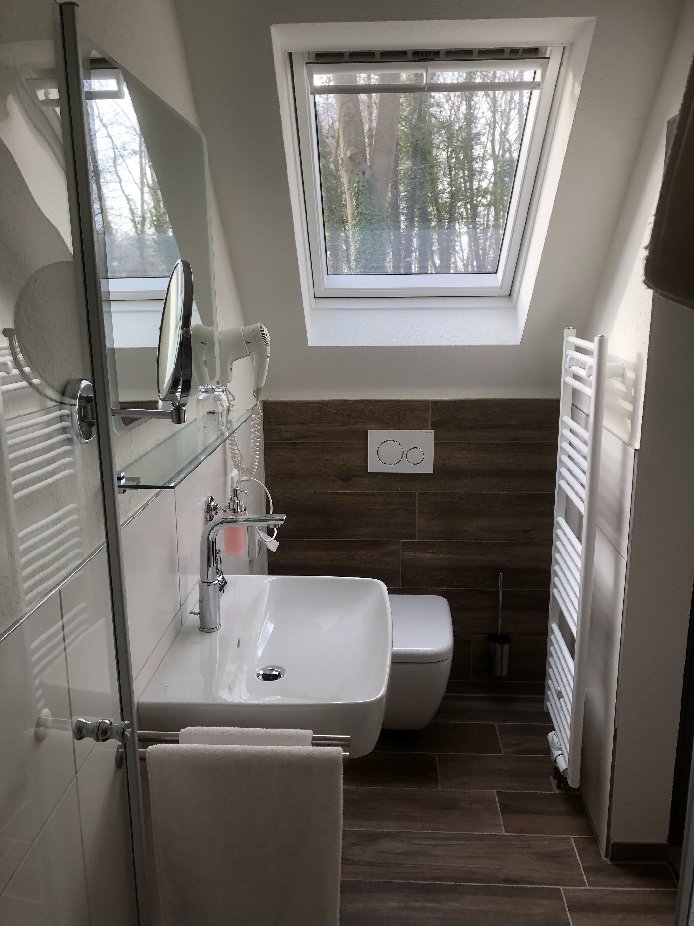 Un baño moderno con cabina de ducha de vidrio, un lavamanos blanco y un inodoro. Hay un tragaluz sobre el inodoro, y una toalla cuelga en la puerta.