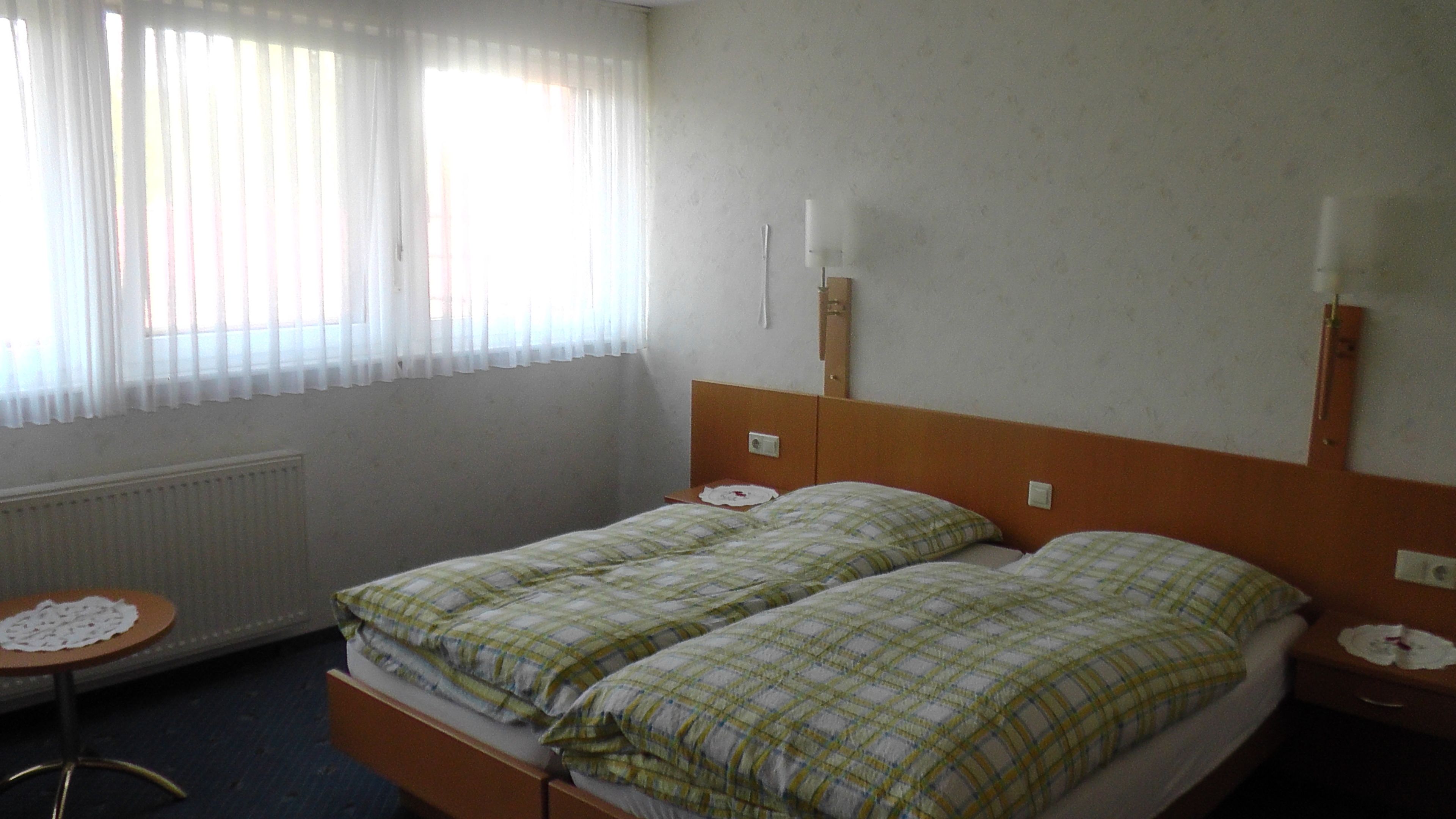 A double bed with a plaid bedspread, a bedside table with a lamp, and a window with sheer curtains.