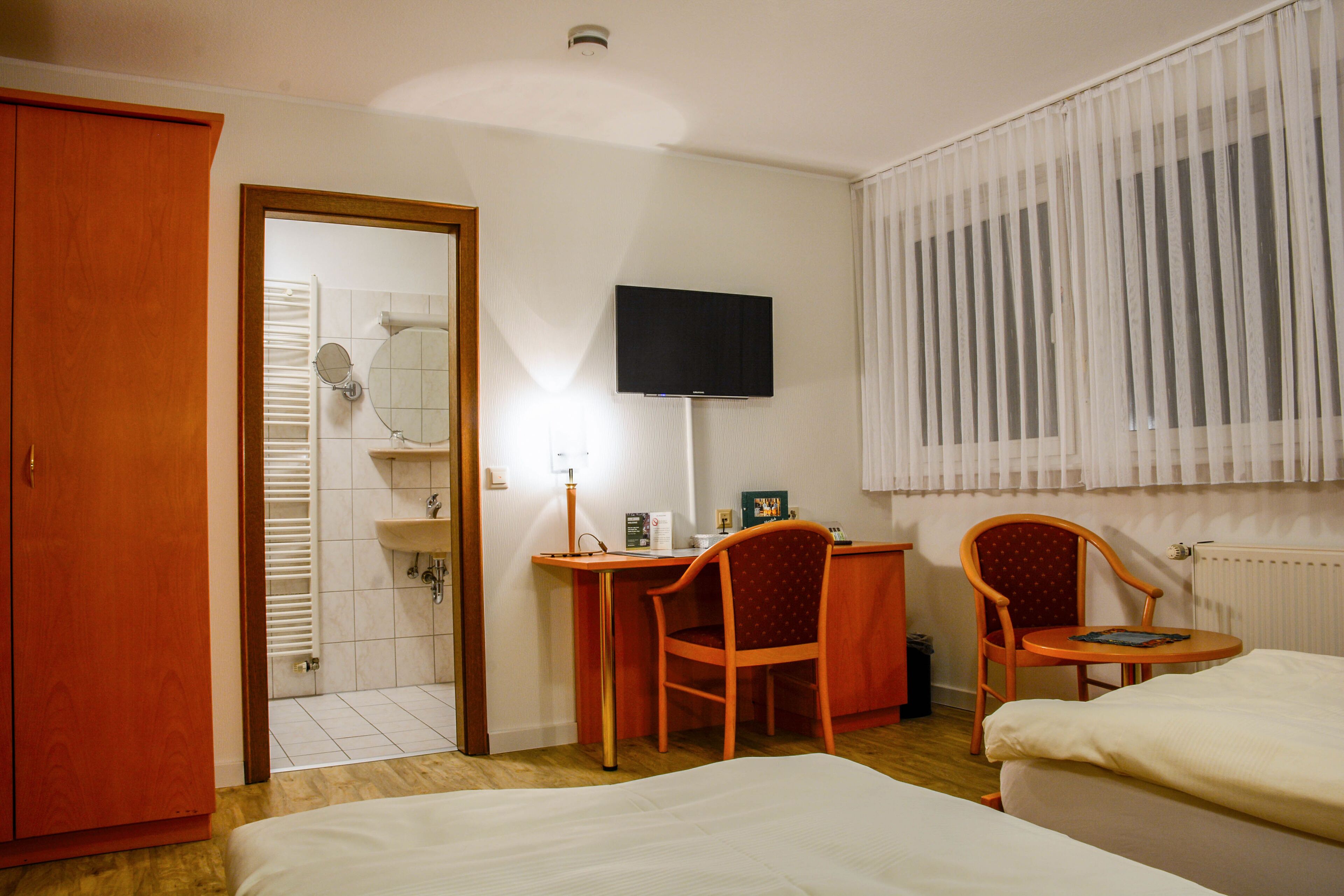 Quarto de hotel com uma cama, duas cadeiras, uma escrivaninha, uma televisão e um banheiro visível por uma porta aberta.