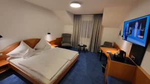 Habitación de hotel con cama, escritorio, silla, televisor y ventana con cortinas.