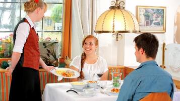 A dining room with a table set for three, a waiter serving a plate of food, and a window with curtains.