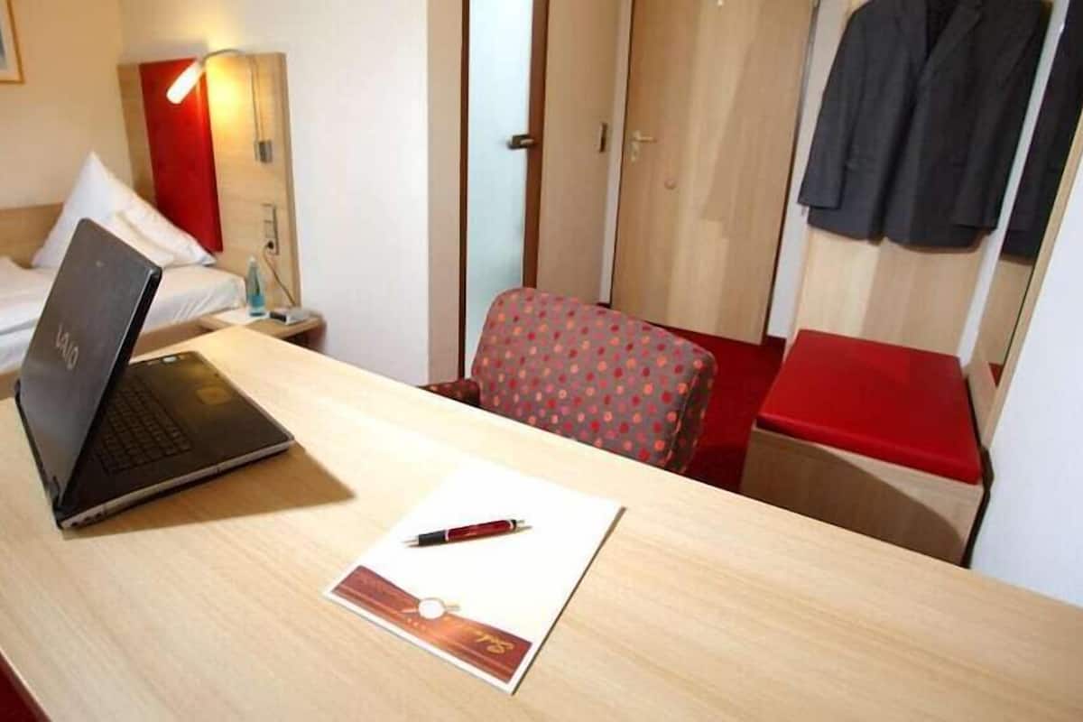 Hotellihuone, jossa on sänky, työpöytä kannettavalla tietokoneella, tuoli, punainen penkki ja naulakko.