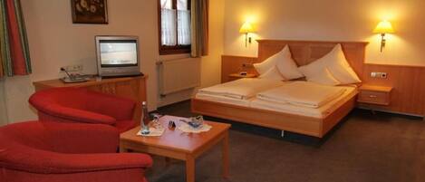 Um quarto de hotel acolhedor com cama, duas luminárias de cabeceira, uma televisão, uma mesinha e uma cadeira vermelha.