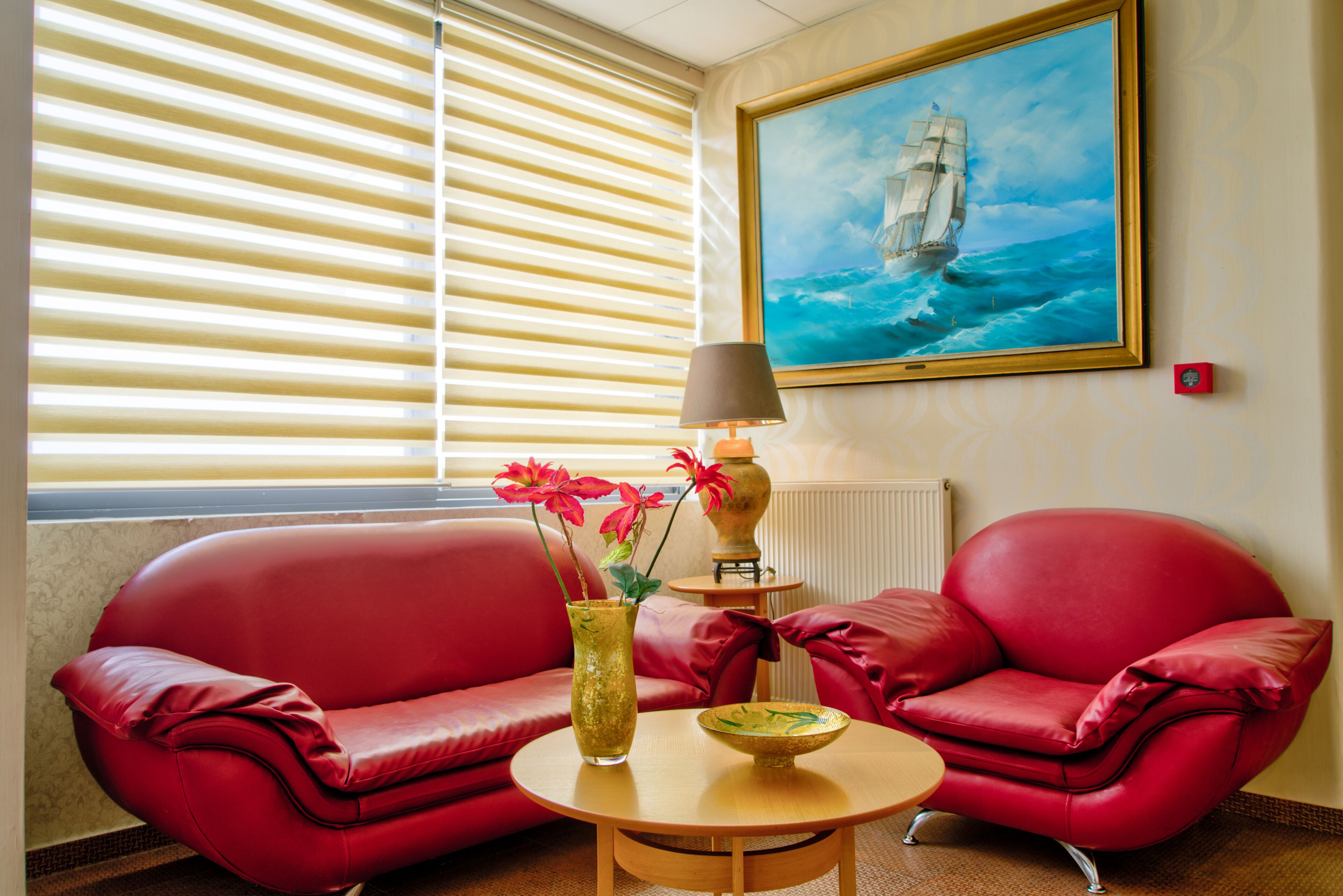 Una habitación con un sofá de cuero rojo, una mesita de madera con un florero y flores, y un cuadro de un barco en la pared.