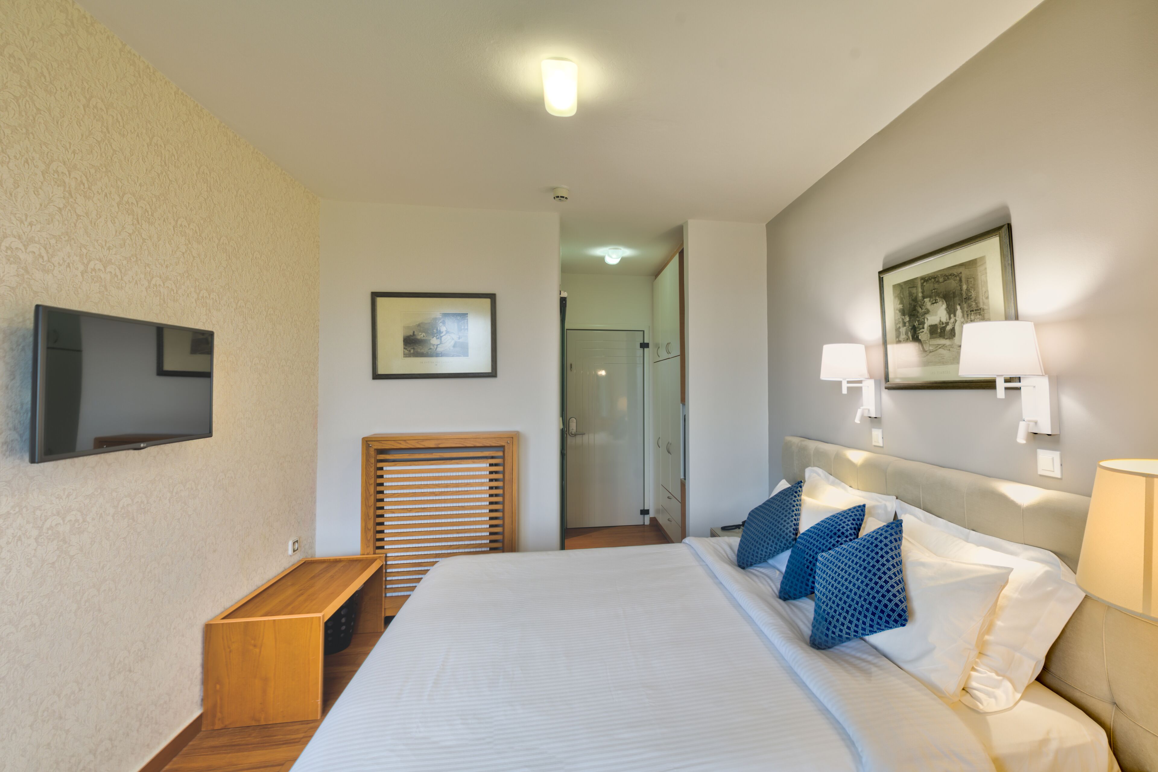 A hotel room with a bed, a TV, a wooden bench, a door, and a wall-mounted picture.