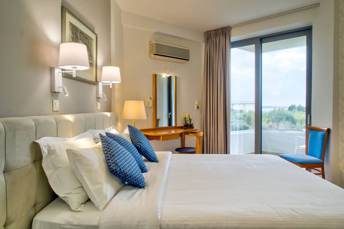 A hotel room with a large bed, a desk with a lamp, a chair, a large window with a view, and an air conditioning unit.