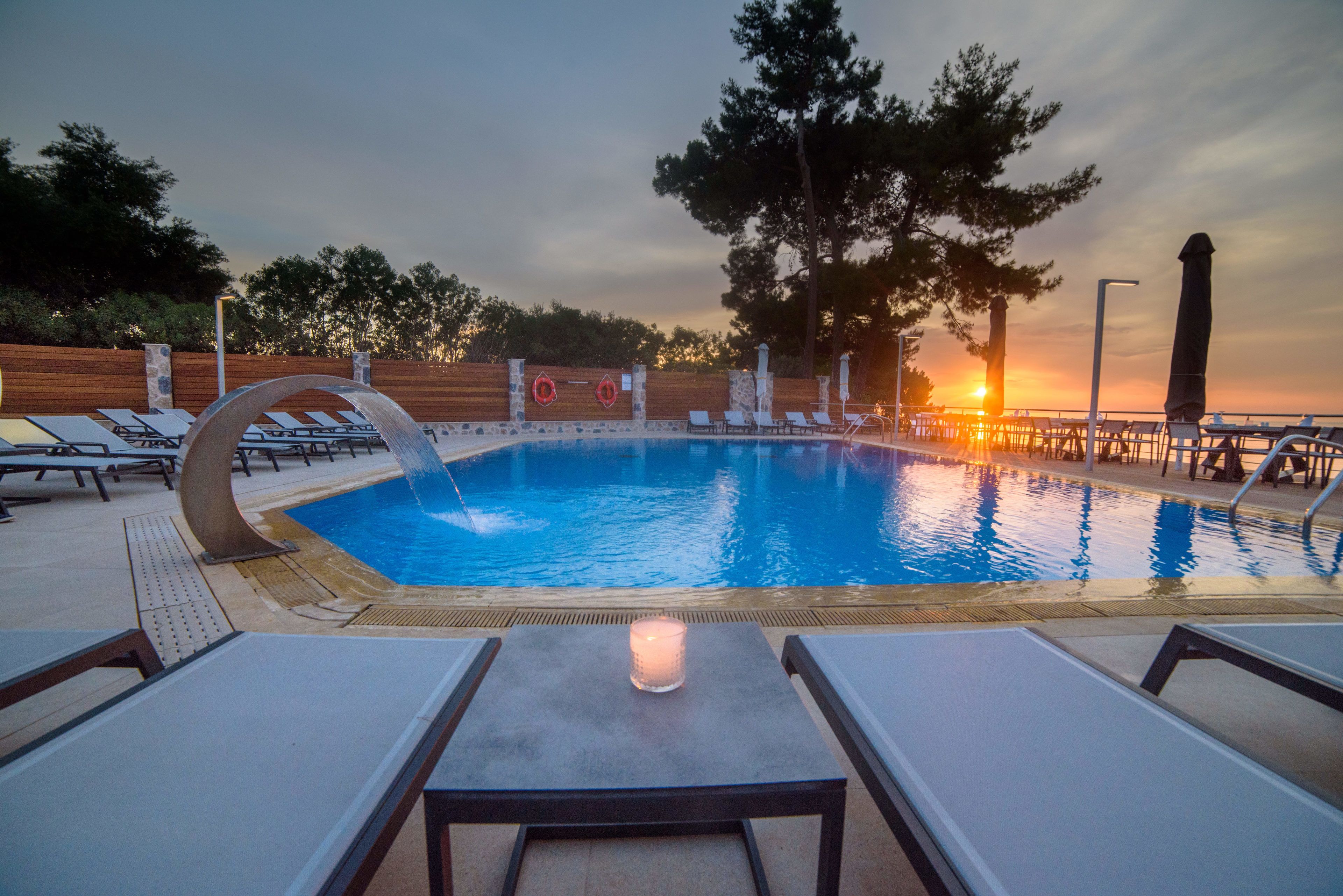 A swimming pool with lounge chairs and a table, surrounded by trees and a sunset in the background.