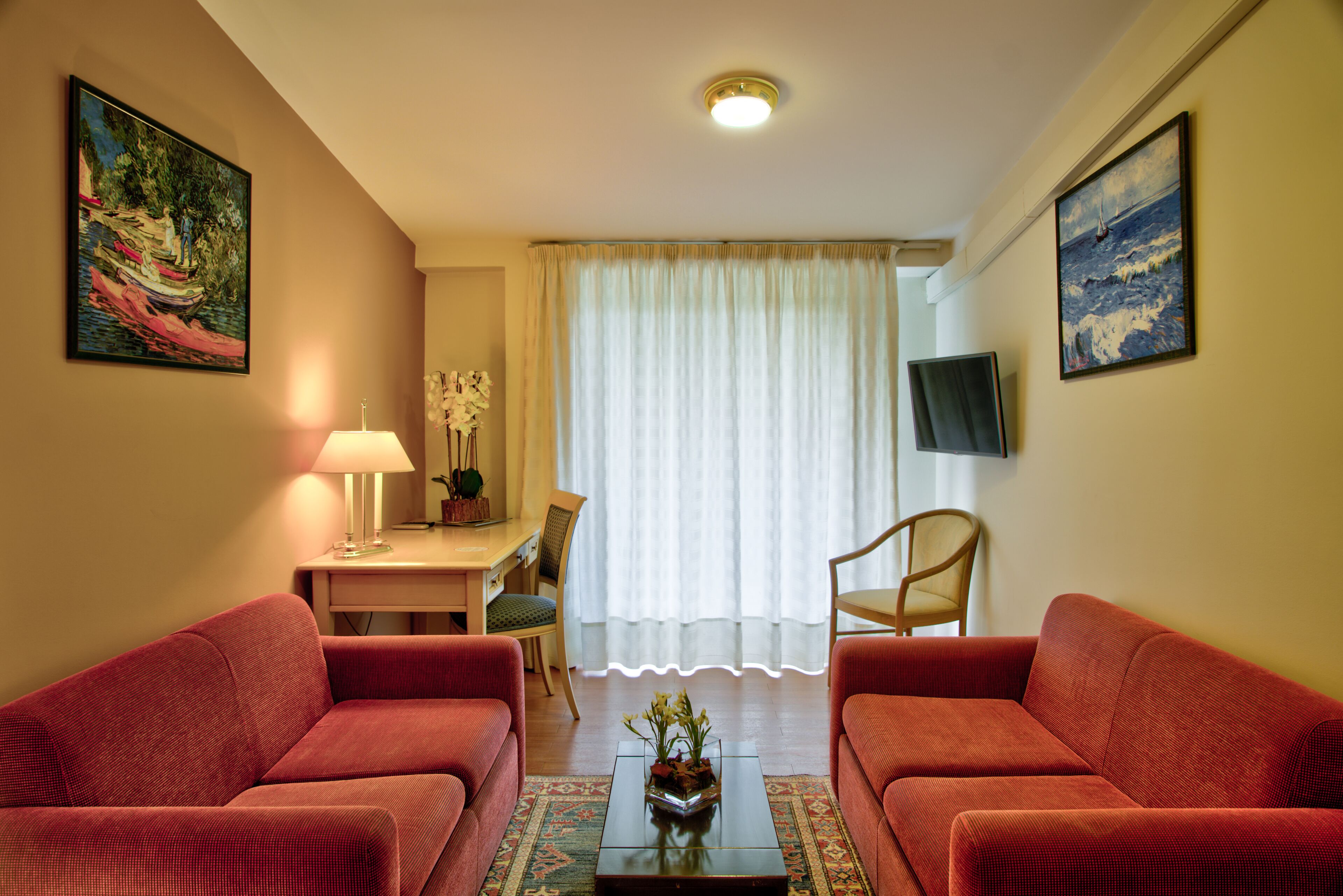 Una sala de estar con dos sofás rojos, una mesa de centro con flores, una lámpara, un televisor en la pared y vistas a una ventana con cortinas.