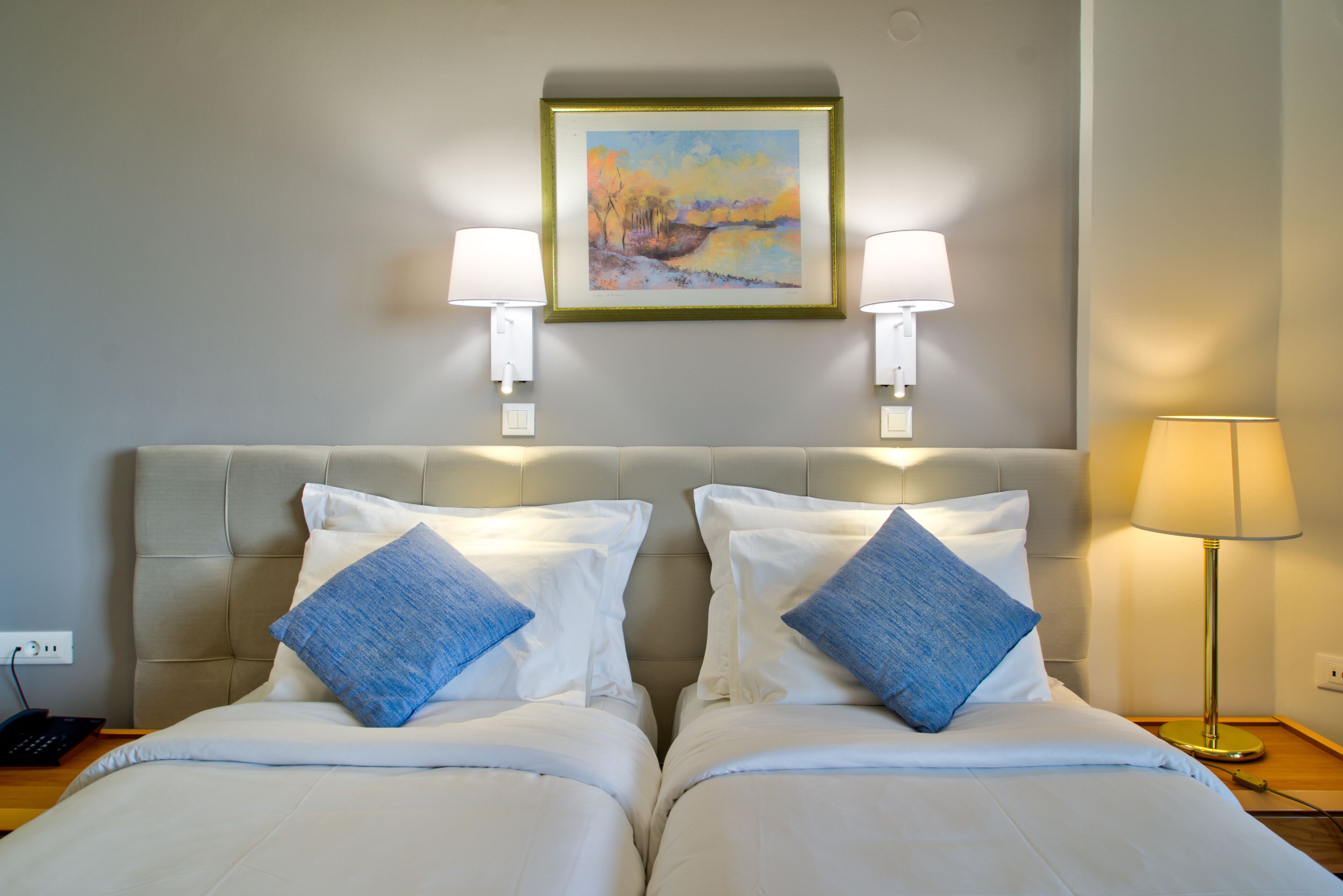 Habitación de hotel con dos camas, ropa de cama blanca, almohadas azules, un cuadro en la pared y dos lámparas de pared.