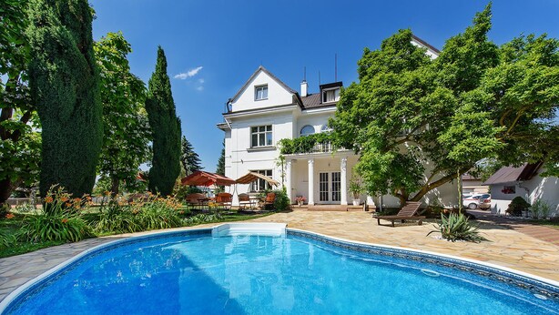 A swimming pool with a tiled deck, surrounded by lush greenery and a white building with a balcony. - Hotel Marie Luisa (Prague)