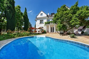 Ein Schwimmbecken mit Fliesenbord, umgeben von üppiger Vegetation und einem weißen Gebäude mit Balkon.