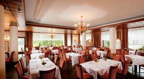 A spacious dining area with multiple tables and chairs, large windows with curtains, and elegant chandeliers. - Parc Hotel Posta (Marebbe)