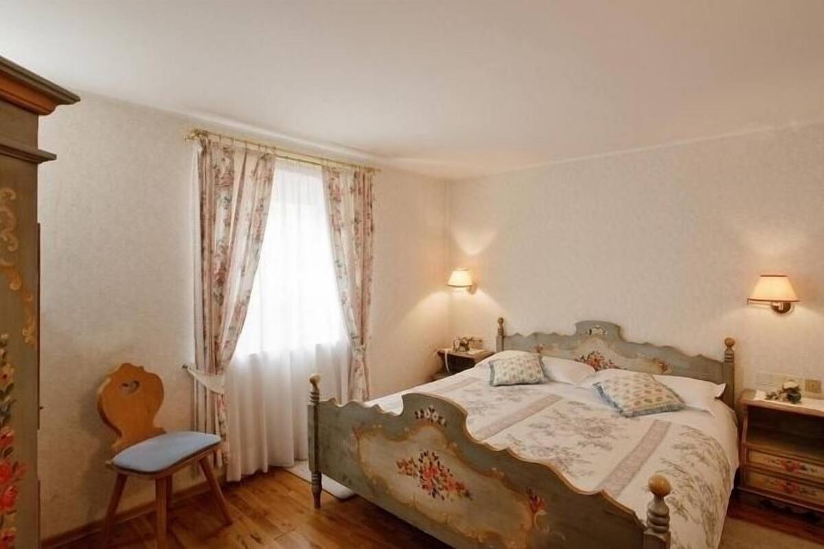 A bedroom with a wooden bed, two bedside tables, a chair, and a window with curtains.