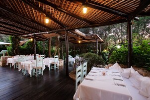 Un área de comedor al aire libre con mesas y sillas, una terraza de madera y una estructura de techo de paja.