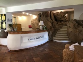 Una mesa de recepción con la leyenda "GREEN PARK HOTEL", un rústico muro de piedra de fondo y una escalera que sube a los pisos superiores.