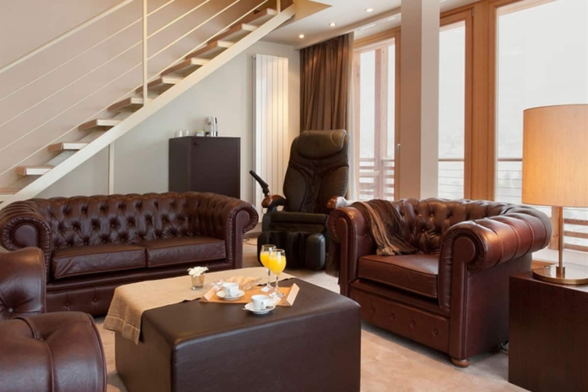 Un vaste salon doté d’un ensemble de meubles en cuir marron, comprenant un canapé et un pouf assortis, ainsi qu’une table basse sur laquelle sont disposés des éléments pour le petit-déjeuner. Une grande fenêtre laisse entrer la lumière naturelle, et un escalier est visible au fond.