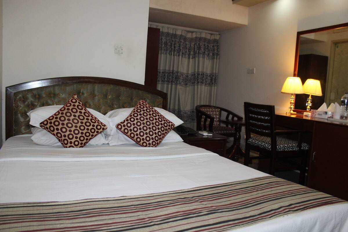 A hotel room with a large bed, two patterned pillows, a striped bedspread, a wooden nightstand, a chair, a desk with a lamp, and a mirror.