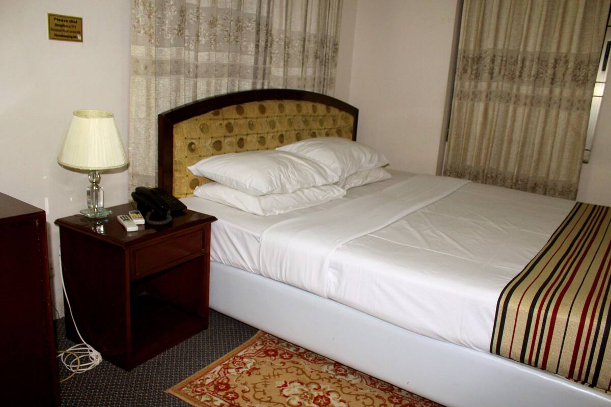 A hotel room with a bed, a nightstand with a lamp, a telephone, and a sign on the wall.