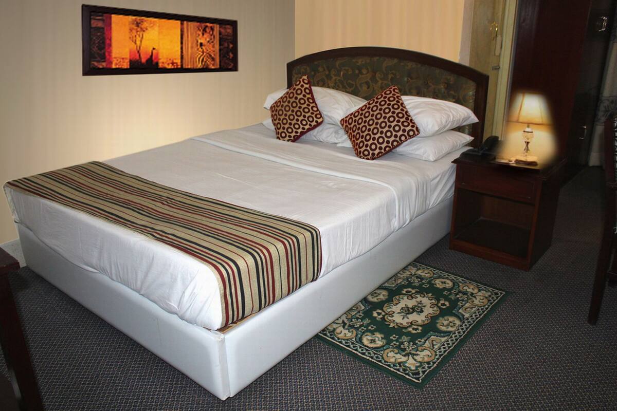 A neatly made bed with a striped blanket, two patterned pillows, a bedside table with a lamp, and a decorative rug.