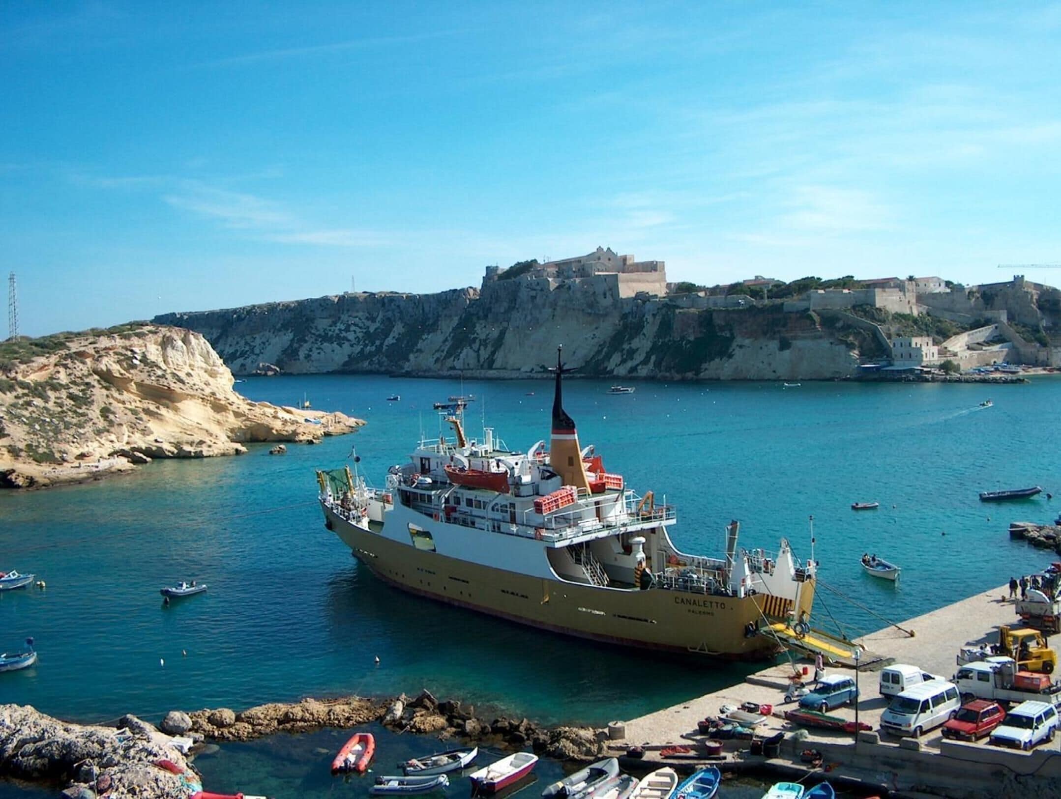 A large ship docked at a port with a historic fort in the background.