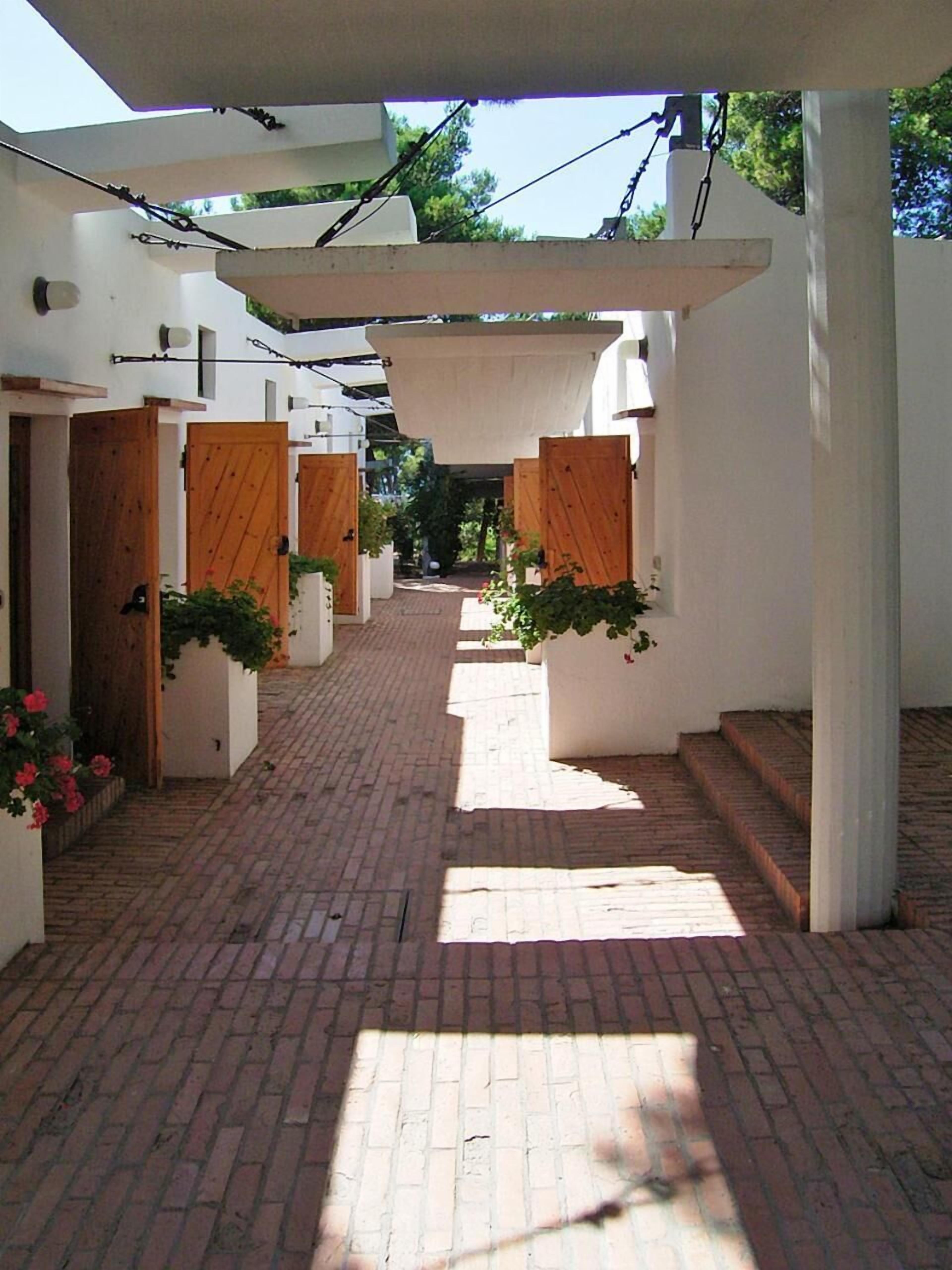 A narrow brick pathway leading to a series of white-walled rooms with wooden doors and potted plants.