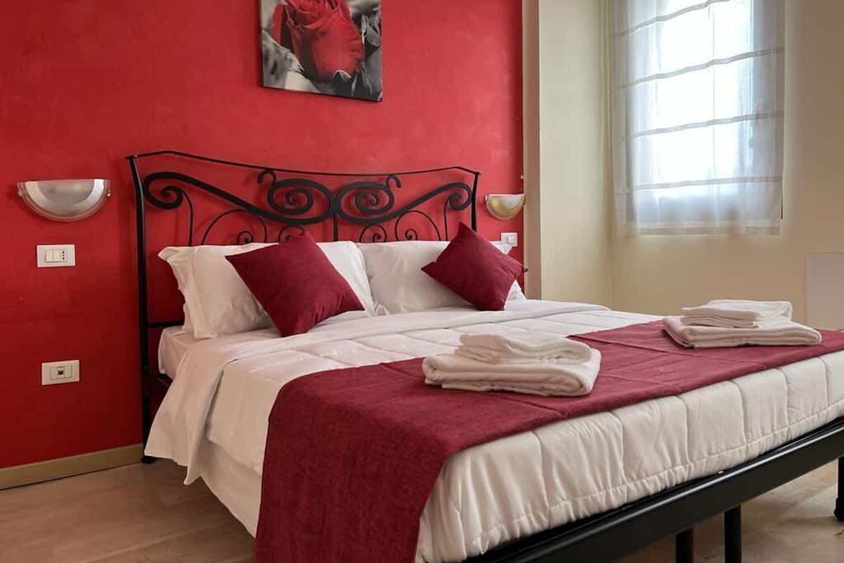 Una cama con ropa de cama blanca y almohadas rojas, una pared roja y una ventana con cortinas traslúcidas.