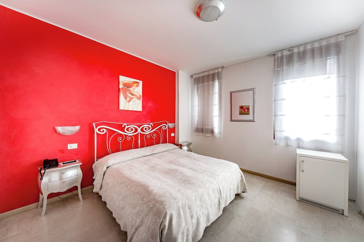 Un dormitorio con una pared roja, una cama blanca, una mesita de noche, un radiador y una ventana con cortinas translúcidas.