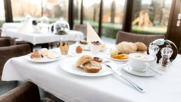 Une table dressée avec une variété d’aliments pour le petit-déjeuner, notamment des viennoiseries, du pain et une tasse de café, avec en arrière-plan une vue sur des arbres et des bâtiments.