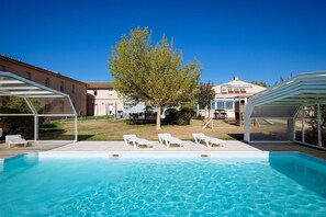 A swimming pool with lounge chairs, a covered seating area, and a building in the background. - Auberge du Pastel (Nailloux)