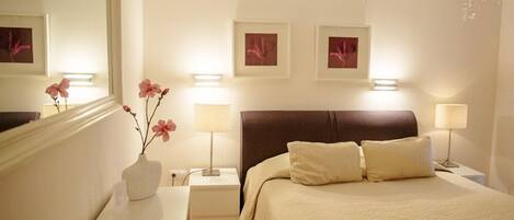 A hotel room with a large bed, two bedside lamps, a nightstand with a vase of flowers, and a chandelier.