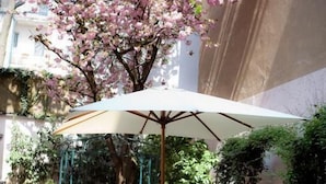 An outdoor seating area with tables and chairs under a large umbrella, surrounded by lush greenery and a blooming tree. - Aurora Hotel (Wiesbaden)