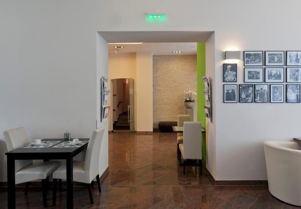 A hotel lobby with a reception desk, a dining area with a table and chairs, a staircase, and a wall with framed pictures.