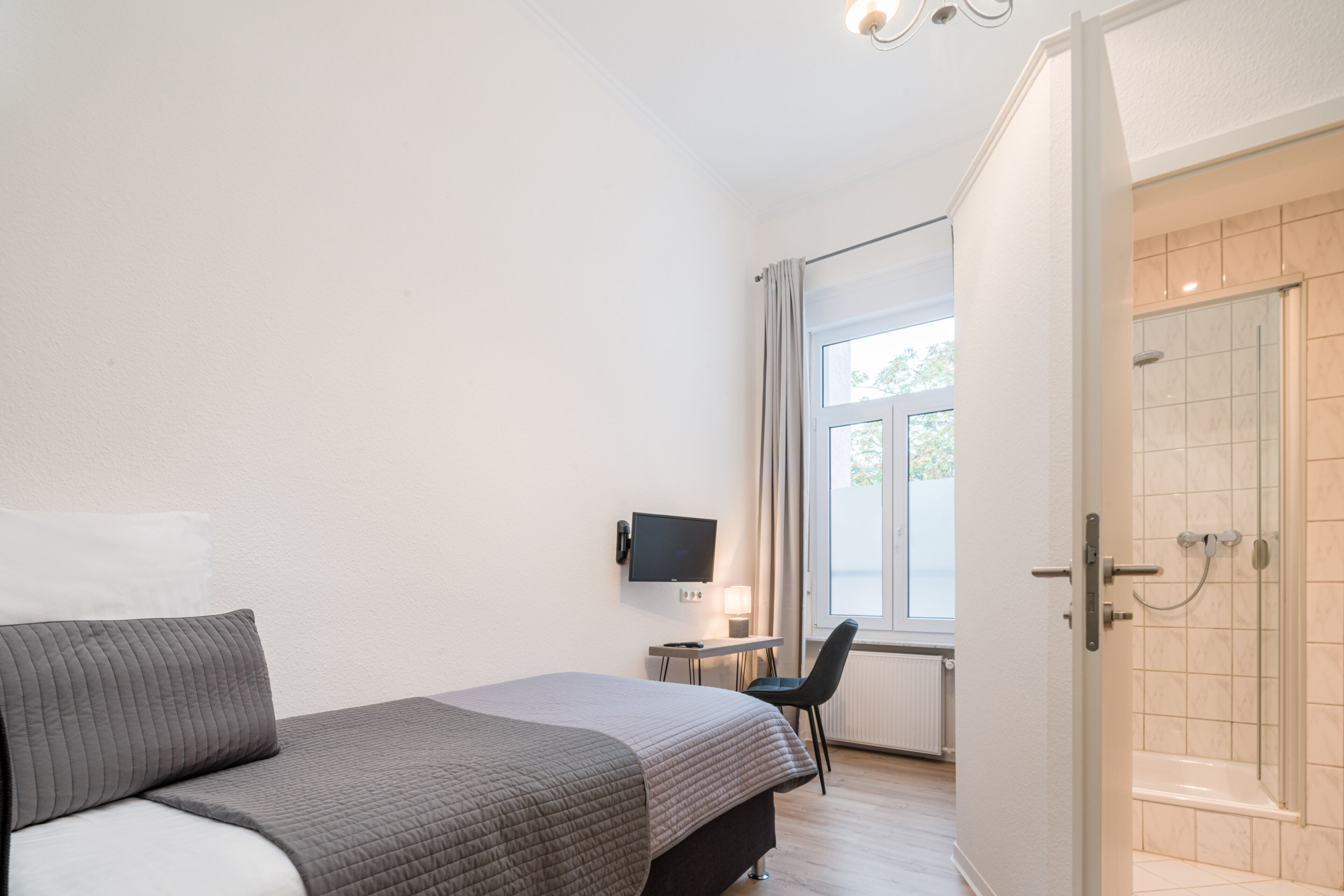 Habitación de hotel con cama, escritorio con silla, televisor y zona de ducha.