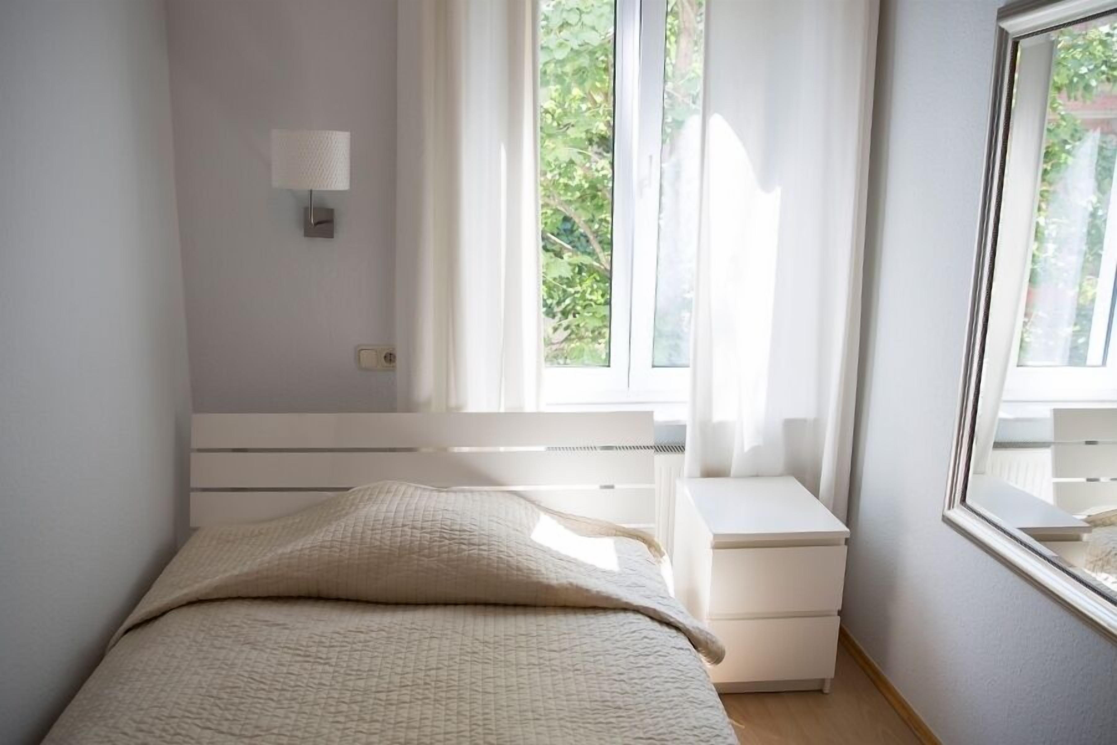 A bedroom with a bed, a nightstand, a wall-mounted lamp, and a window with curtains.