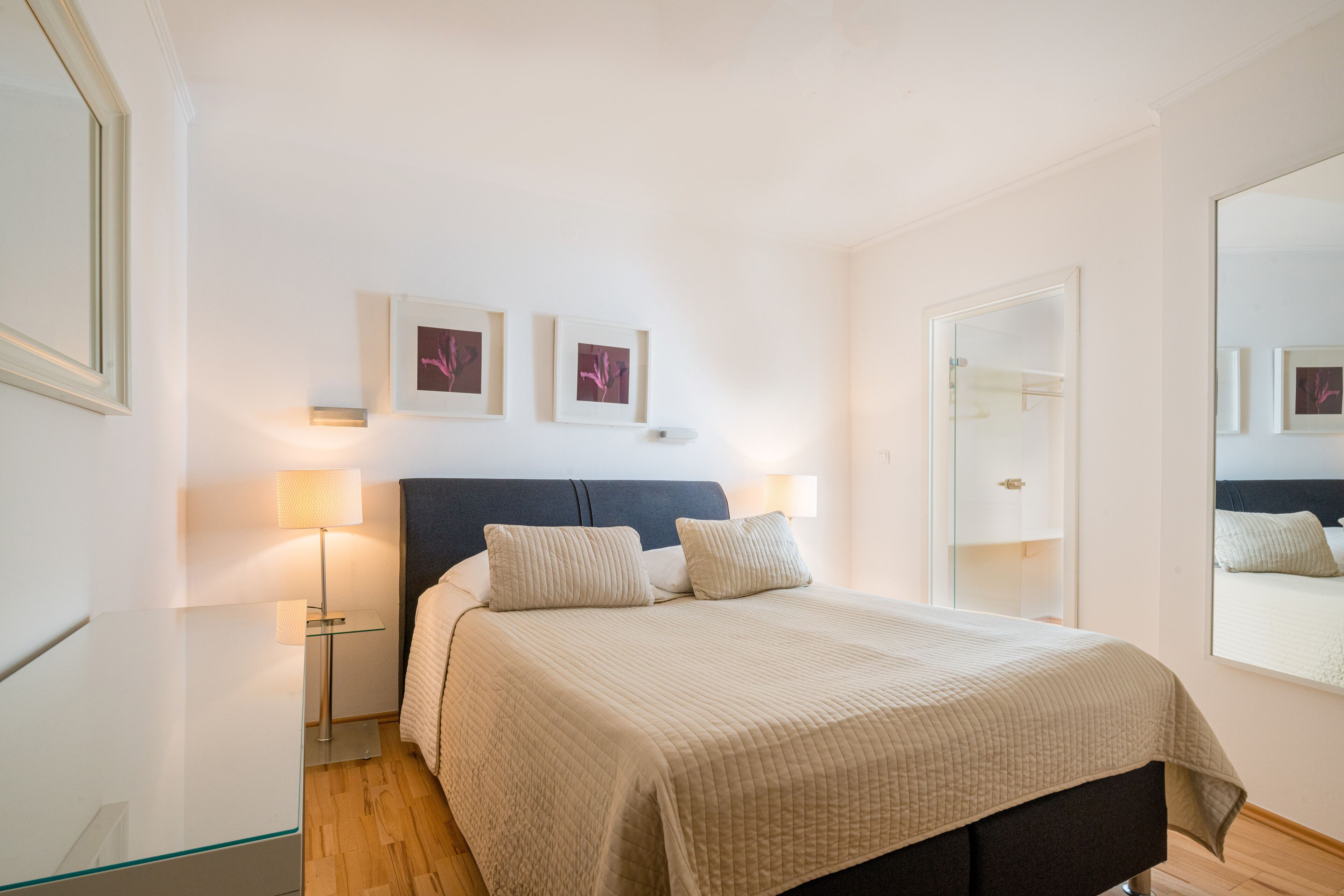 A bedroom with a bed, bedside lamps, a nightstand, a mirror, and framed pictures on the wall.