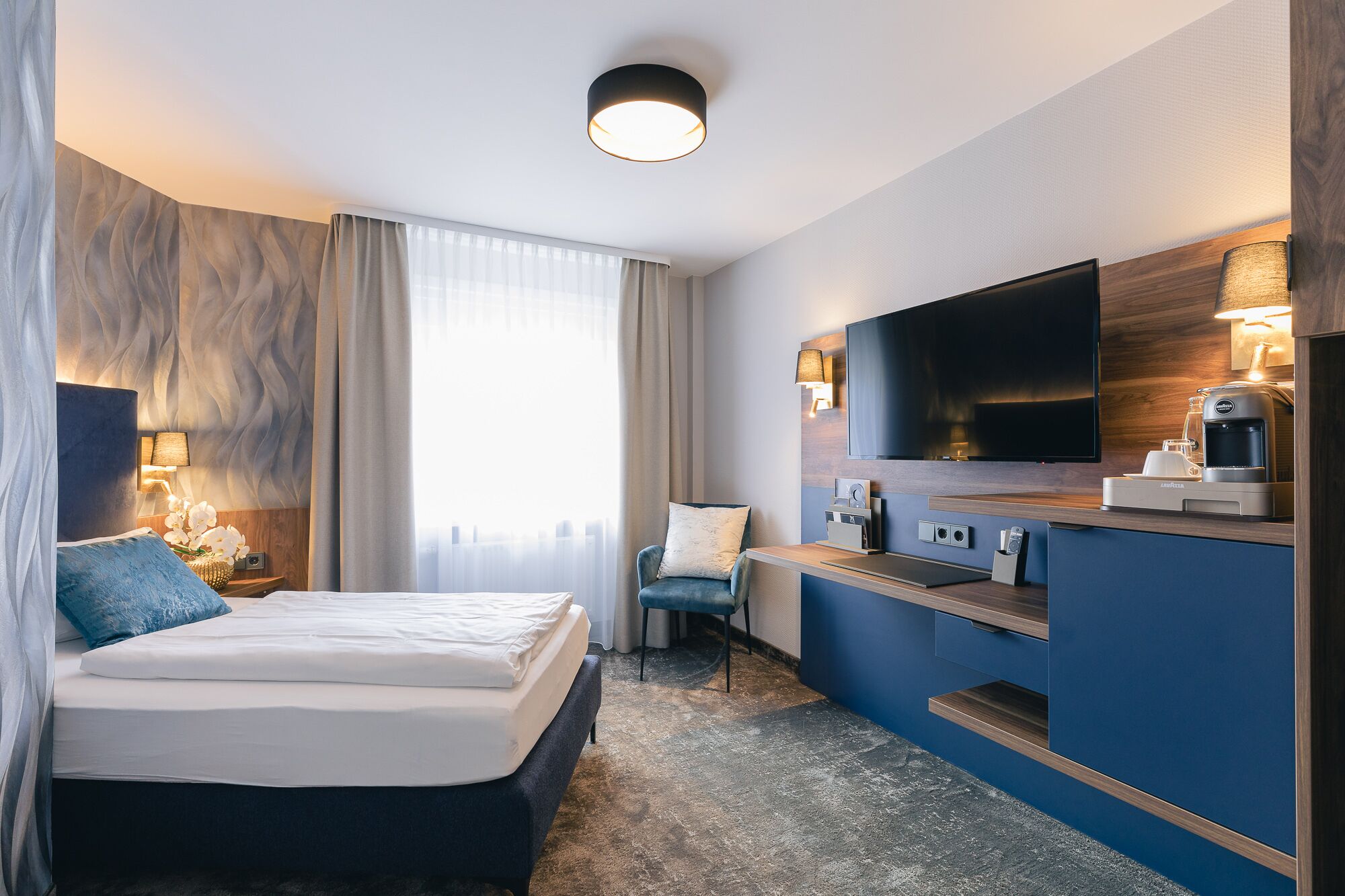 A hotel room with a bed, a desk with a flat-screen TV, a coffee machine, and a chair.