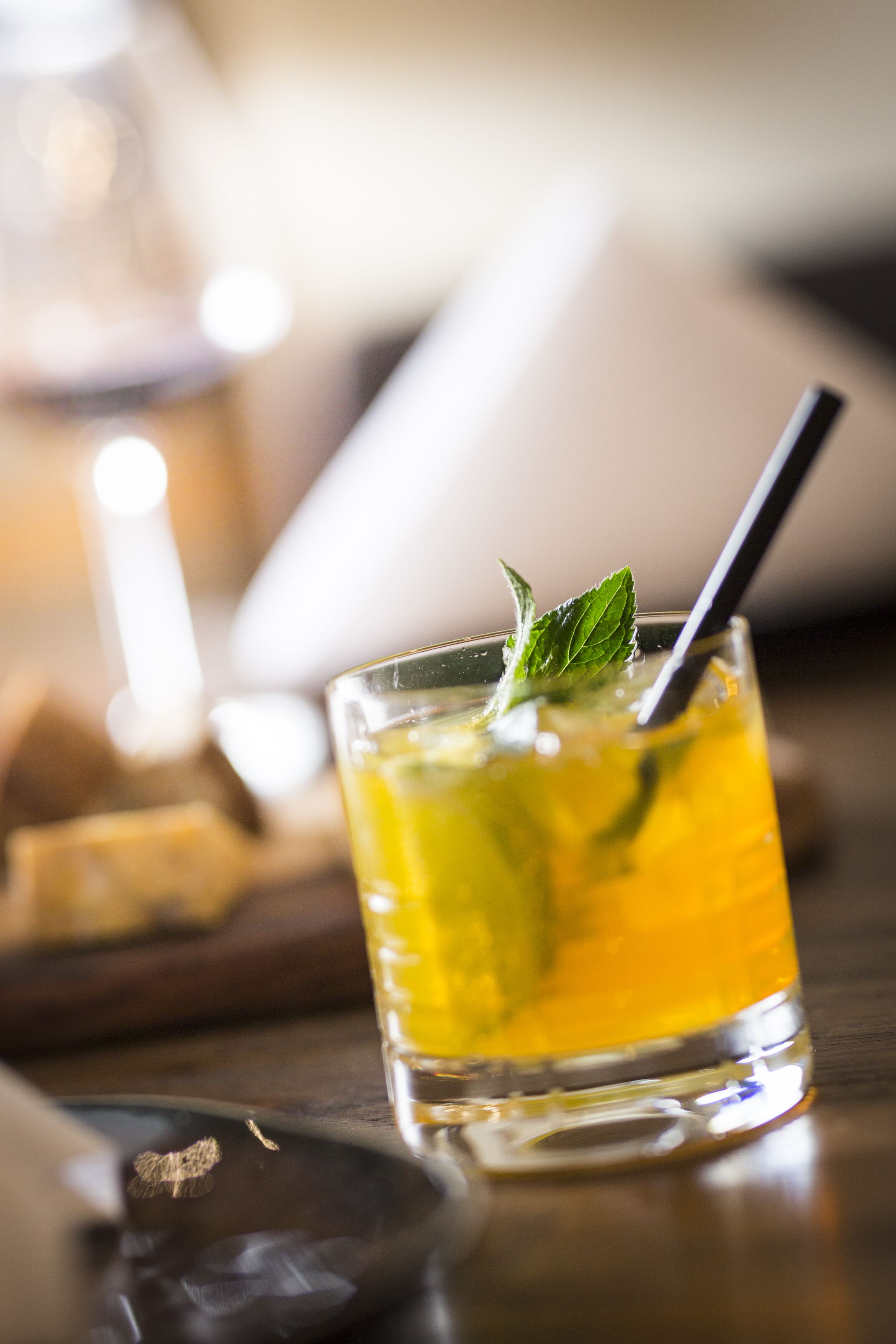 A glass of yellow beverage with a mint garnish and a straw, placed on a wooden surface.