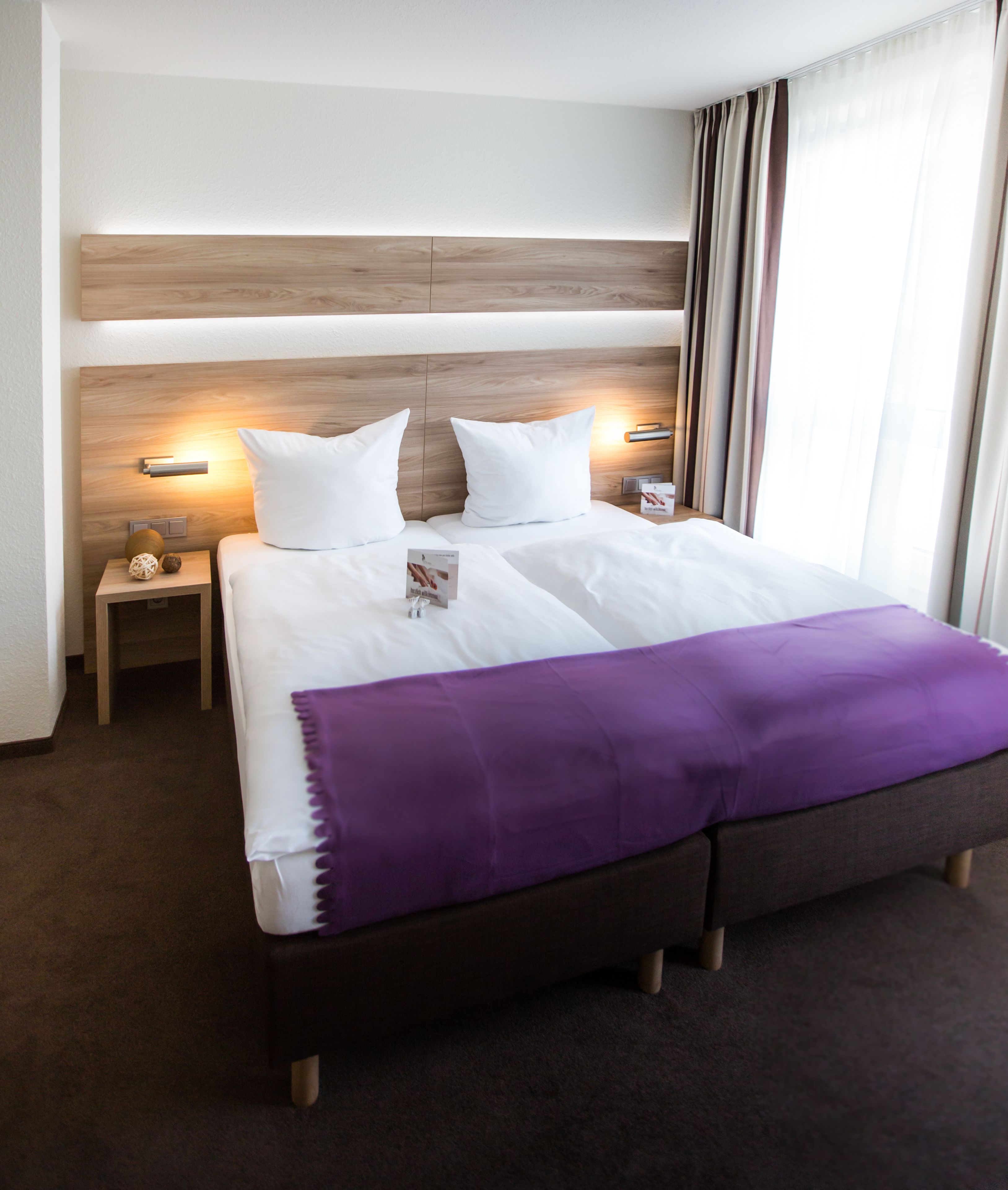 A hotel room with a large bed, two bedside tables, a wooden headboard, and a window with curtains.