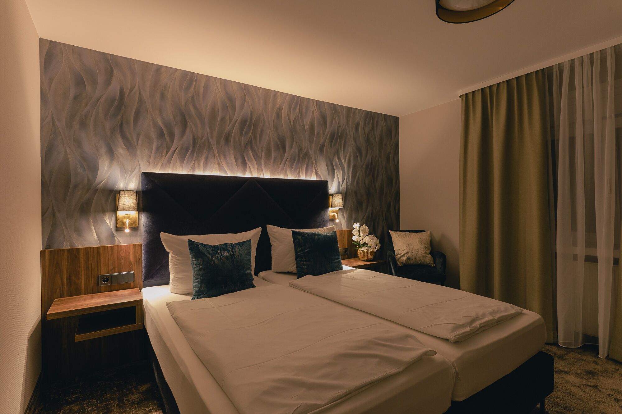 A hotel room with a large bed, two bedside tables, a wall-mounted lamp, and a window with curtains.