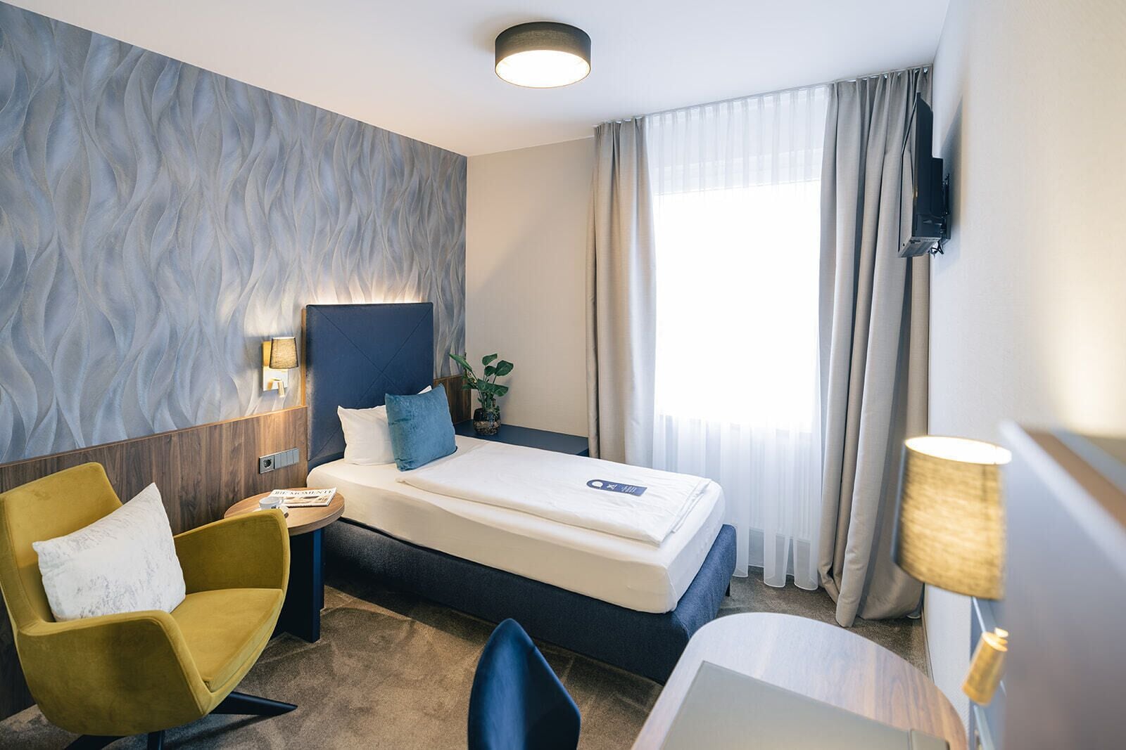 Una habitación de hotel con cama, un sillón amarillo, escritorio, lámpara y televisor.